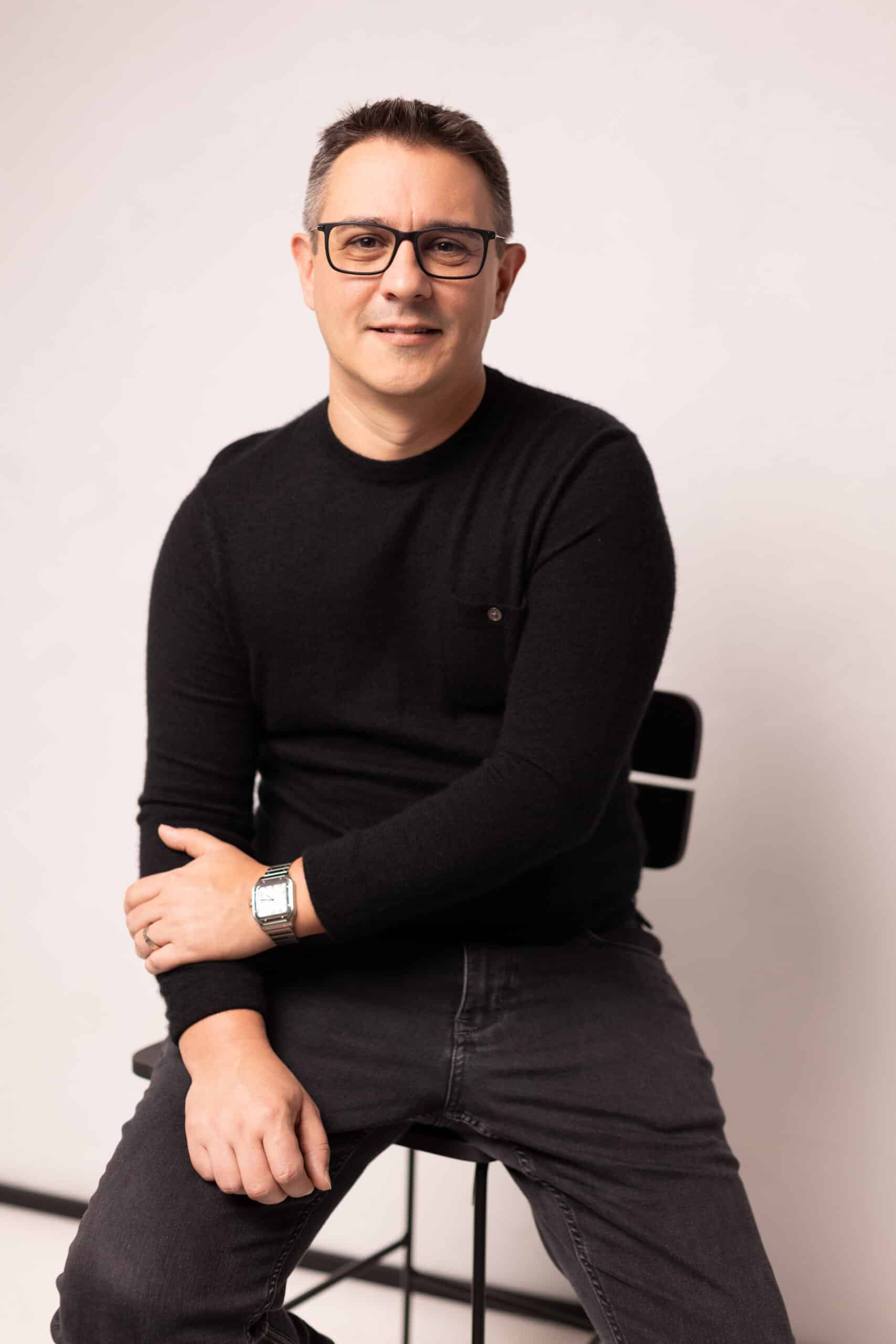 Man wearing glasses, a black sweater, and dark jeans sitting on a stool against a plain background.