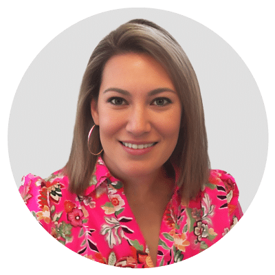 Woman with shoulder-length light brown hair, wearing a pink floral blouse and hoop earrings, smiling against a light gray background.