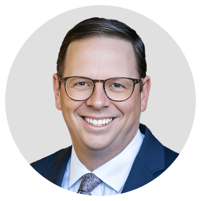 A man in a suit and tie smiling at the camera, wearing glasses.