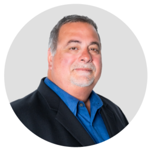 A man wearing a black blazer and blue shirt, smiling with a neutral background.