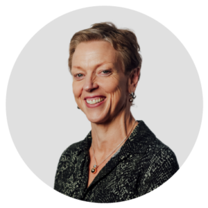 Smiling person with short hair wearing a dark patterned shirt and jewelry, against a light gray background.