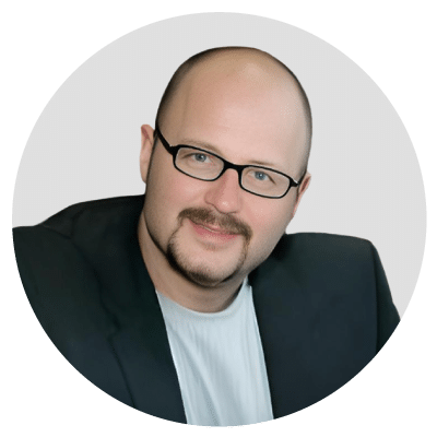 A person with glasses and a goatee wearing a black blazer and white shirt, smiling.