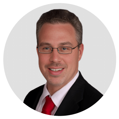 A man wearing glasses and a suit with a red tie, smiling against a neutral background.