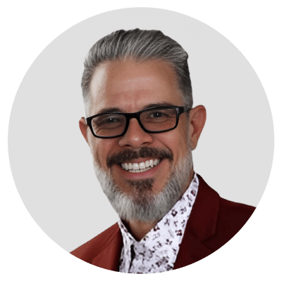 Man with glasses, gray hair, and beard smiling, wearing a patterned shirt and maroon blazer.