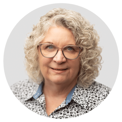 A woman with curly gray hair, wearing glasses and a patterned blouse, against a light gray background.