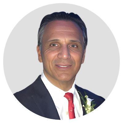 A man wearing a dark suit with a red tie and a white flower boutonniere against a neutral background.
