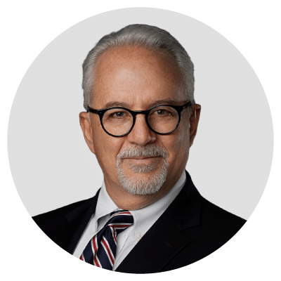 A man with gray hair and a beard wearing glasses, a suit, and a striped tie.