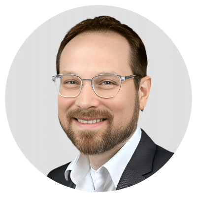 A man with glasses and a beard wearing a suit and white shirt, smiling against a light gray background. A man with glasses and a beard wearing a suit and white shirt, smiling against a light gray background.
