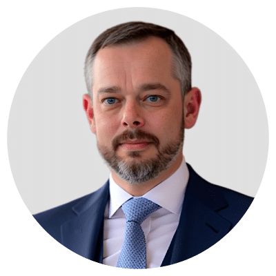 A man with short hair and a beard, wearing a suit and tie, against a light gray background.
