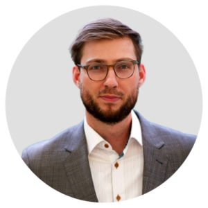 A man with glasses and a beard wearing a gray suit and white shirt against a plain background.