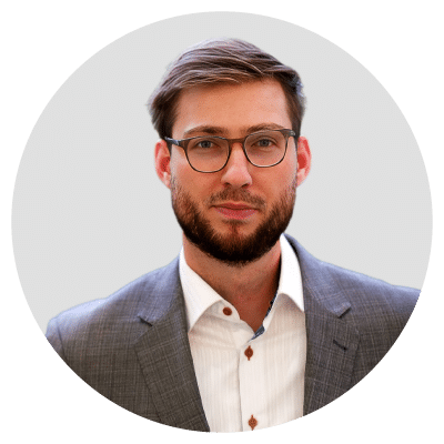 A man with glasses and a beard wearing a gray suit and white shirt against a plain background.