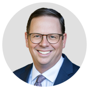 A smiling man wearing glasses, a suit, and a tie.