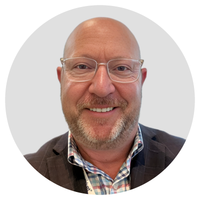 A bald man with glasses and a beard, wearing a checkered shirt and a dark blazer, smiling at the camera.